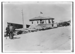 Ön planda yürüyen iki kişi ve çatısında Türk bayrağı bulunan tarihî bir bina.