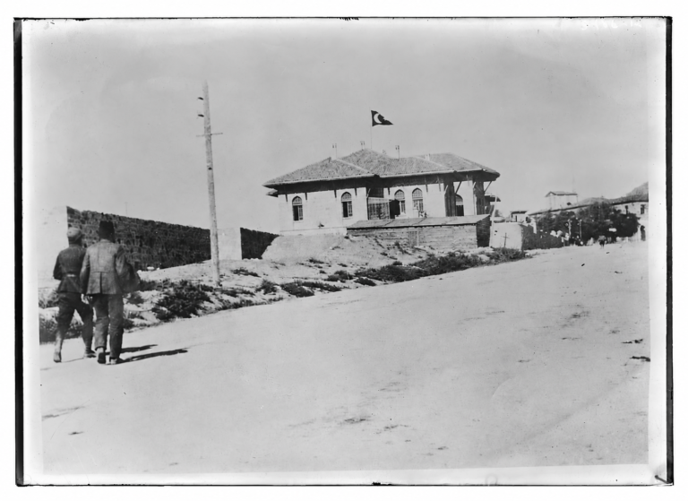 Ön planda yürüyen iki kişi ve çatısında Türk bayrağı bulunan tarihî bir bina.