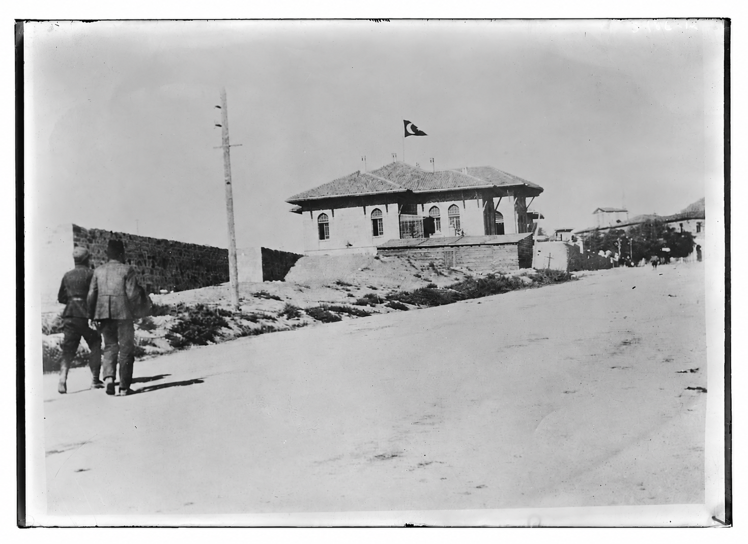Ön planda yürüyen iki kişi ve çatısında Türk bayrağı bulunan tarihî bir bina.